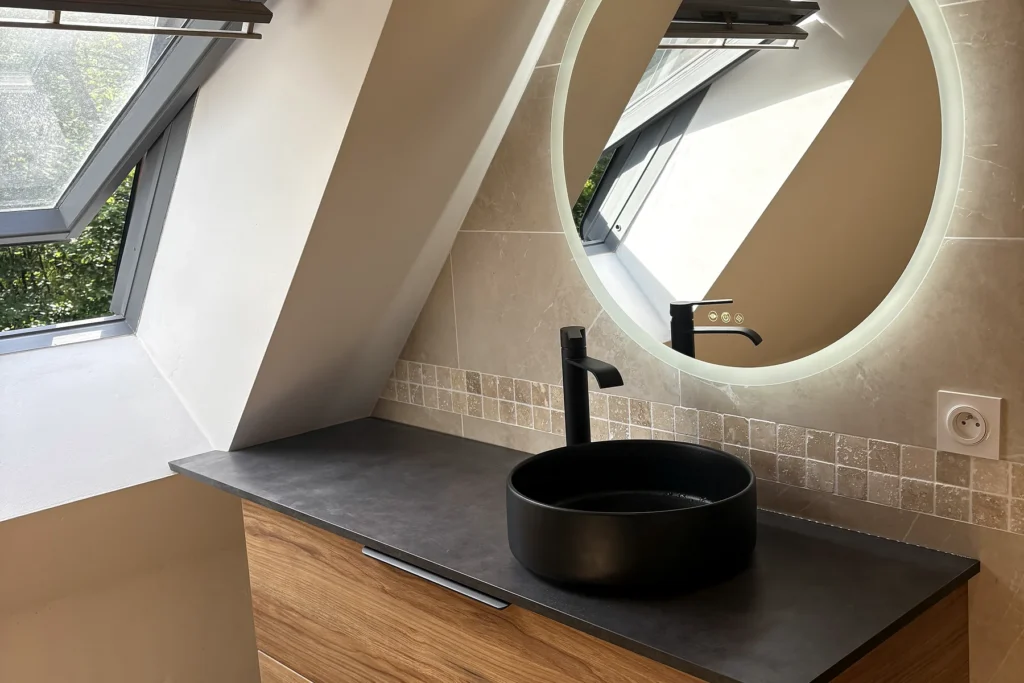 Meuble vasque en bois avec vasque noire, miroir rond lumineux et faïence beige.