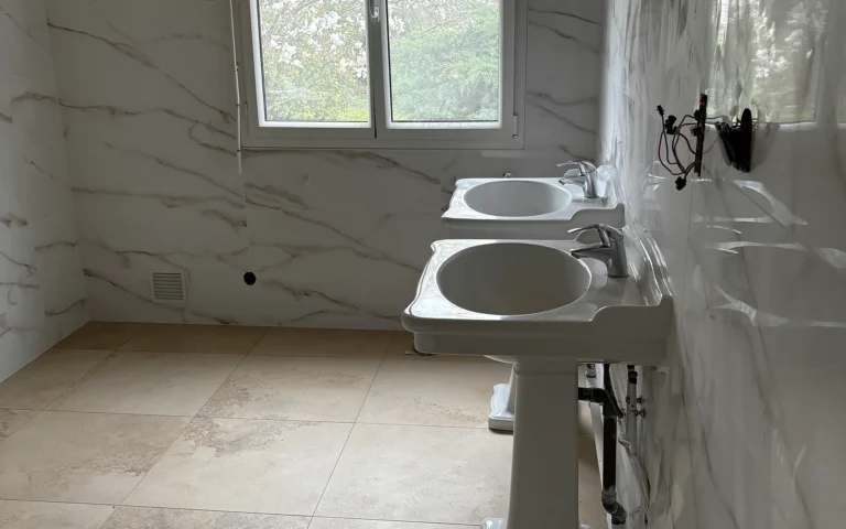 Salle de bains rénovée à L’Hermitage avec double vasque, WC et faïence effet marbre.