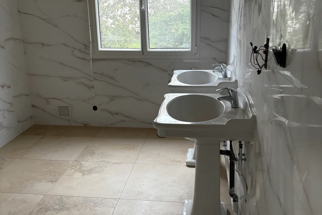 Salle de bains rénovée avec double vasque, WC, fenêtre et faïence effet marbre.