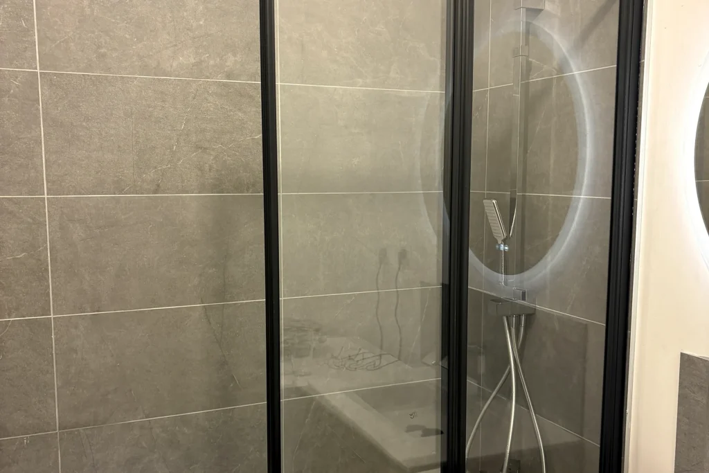 Salle de bains après rénovation avec nouvelle baignoire, paroi vitrée noire et faïence grise.