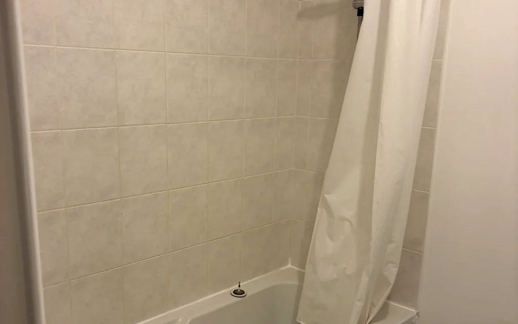 Salle de bains avant rénovation avec ancienne baignoire, rideau de douche et faïence claire.