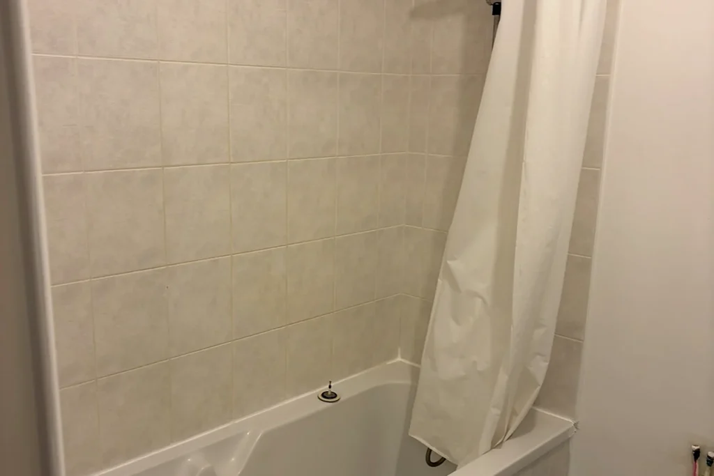 Salle de bains avant rénovation avec ancienne baignoire, rideau de douche et faïence claire.