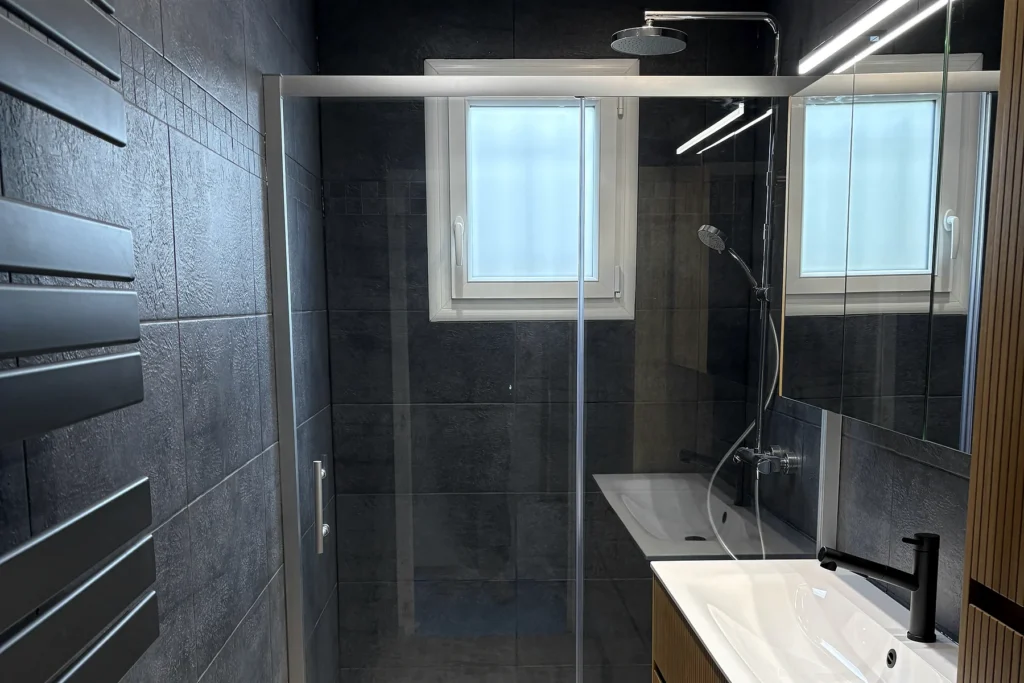 Salle de bains rénovée avec douche vitrée, meuble vasque en bois et faïence sombre.