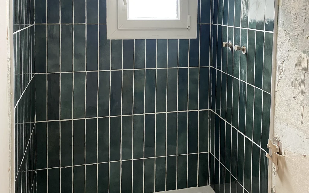 Espace douche rénové avec receveur blanc et faïence murale vert foncé autour de la fenêtre.