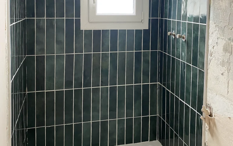 Espace douche rénové avec receveur blanc et faïence murale vert foncé autour de la fenêtre.