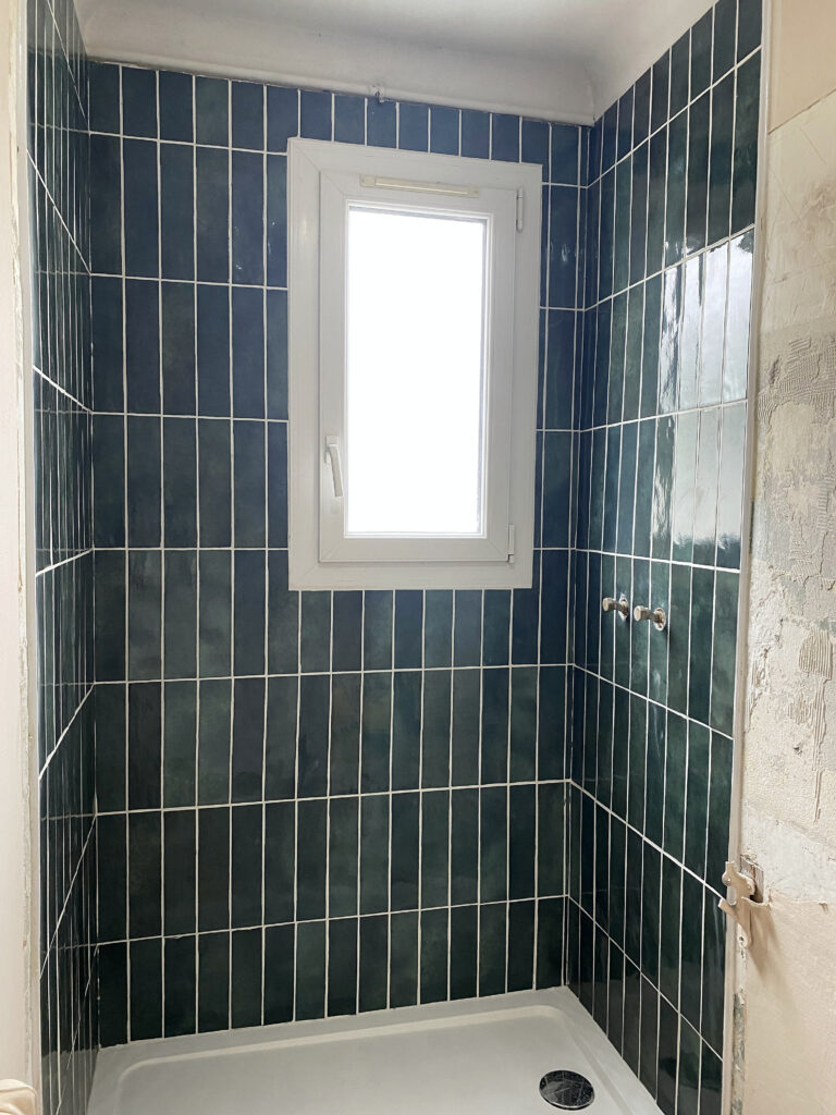 Espace douche rénové avec receveur blanc et faïence murale vert foncé autour de la fenêtre.
