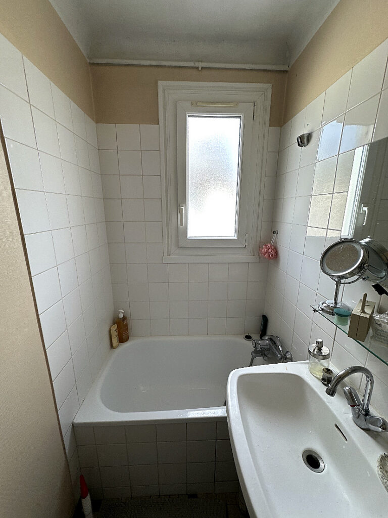Salle de bains avant rénovation avec baignoire, faïence blanche, lavabo et fenêtre.
